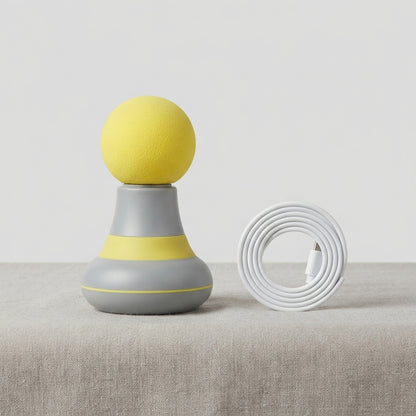 A modern desk lamp with a yellow spherical top, a gray and yellow textured base, and a coiled white charging cable placed beside it on a light beige fabric surface against a neutral light gray background.